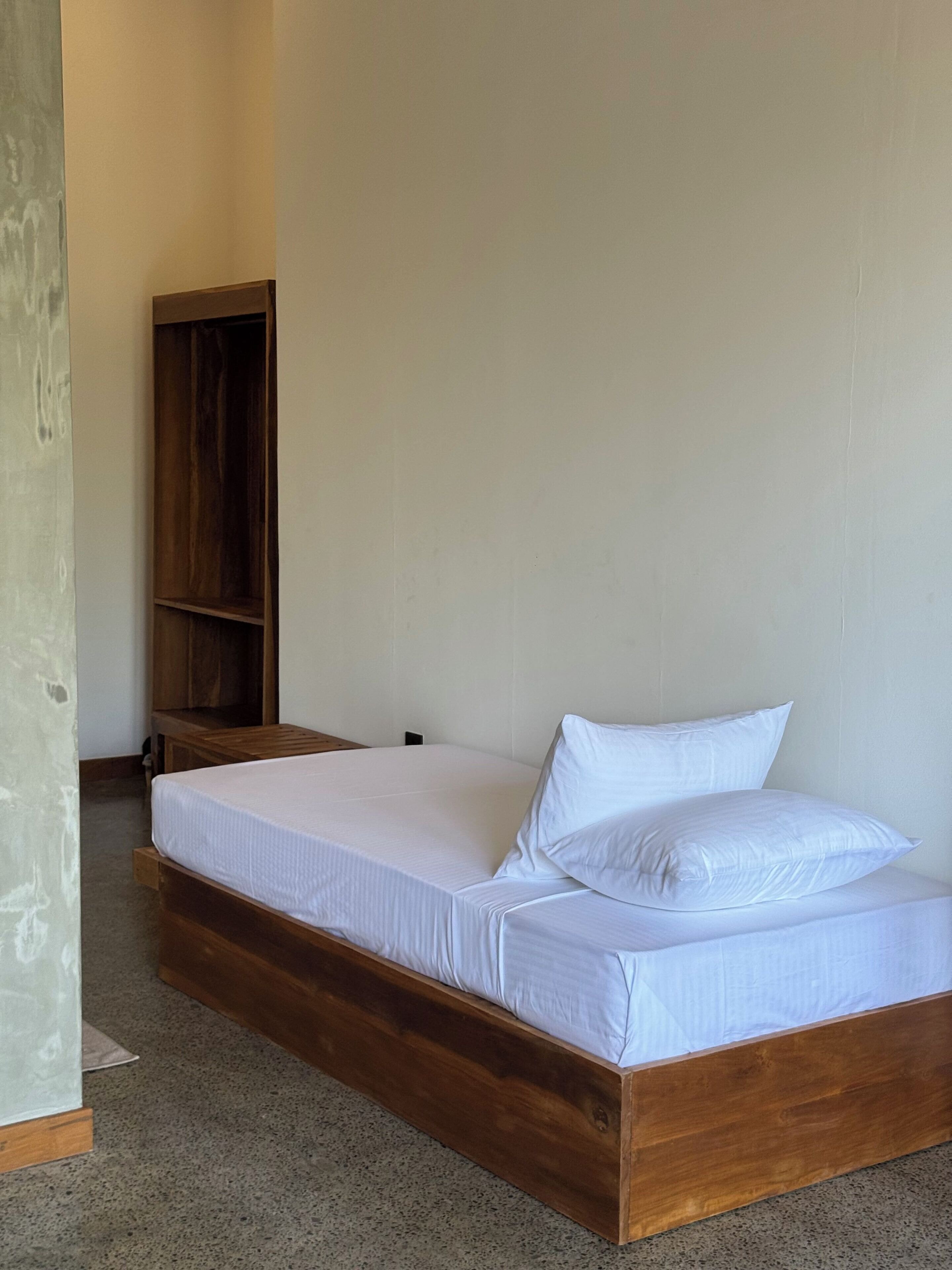 Un lit simple avec une literie blanche, dans une chambre dotée d’une tête de lit et d’une armoire en bois.