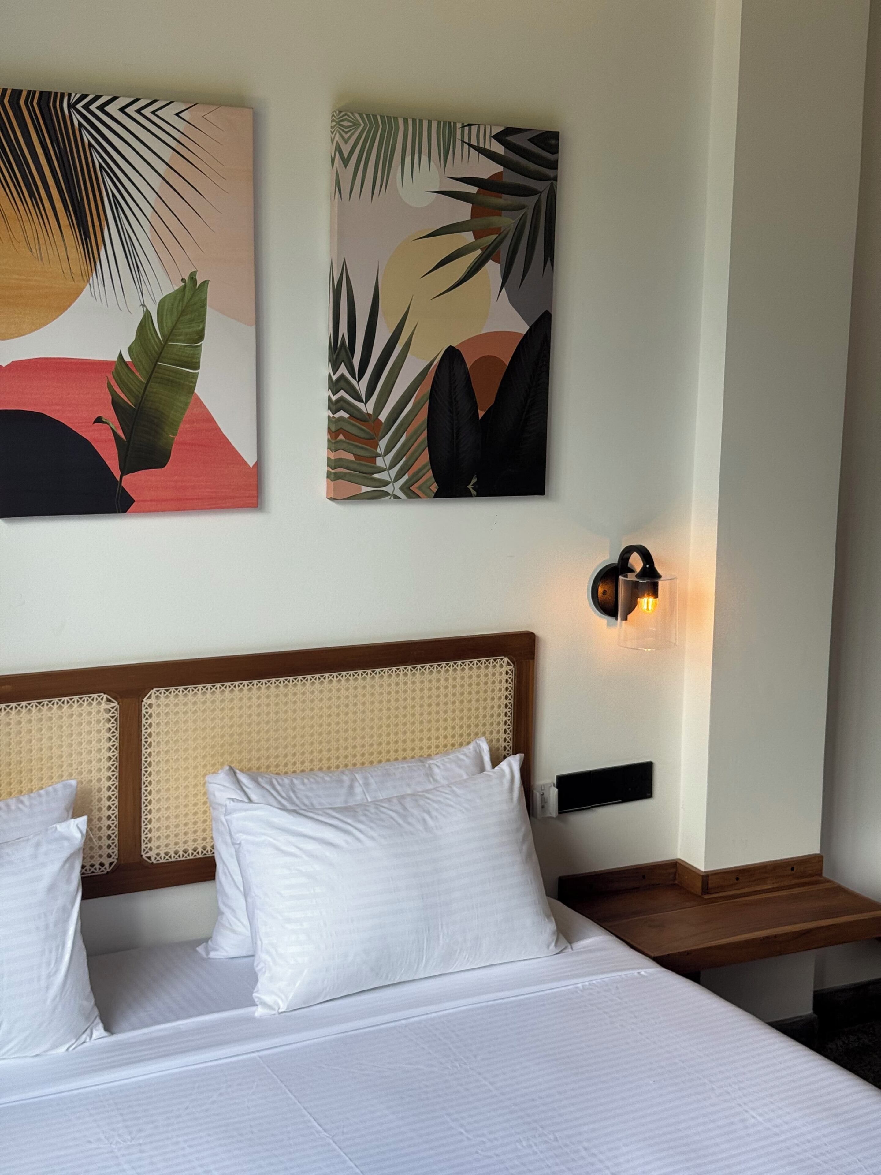 A hotel room with a bed, white pillows, a headboard, a wall-mounted light, and two pieces of wall art.