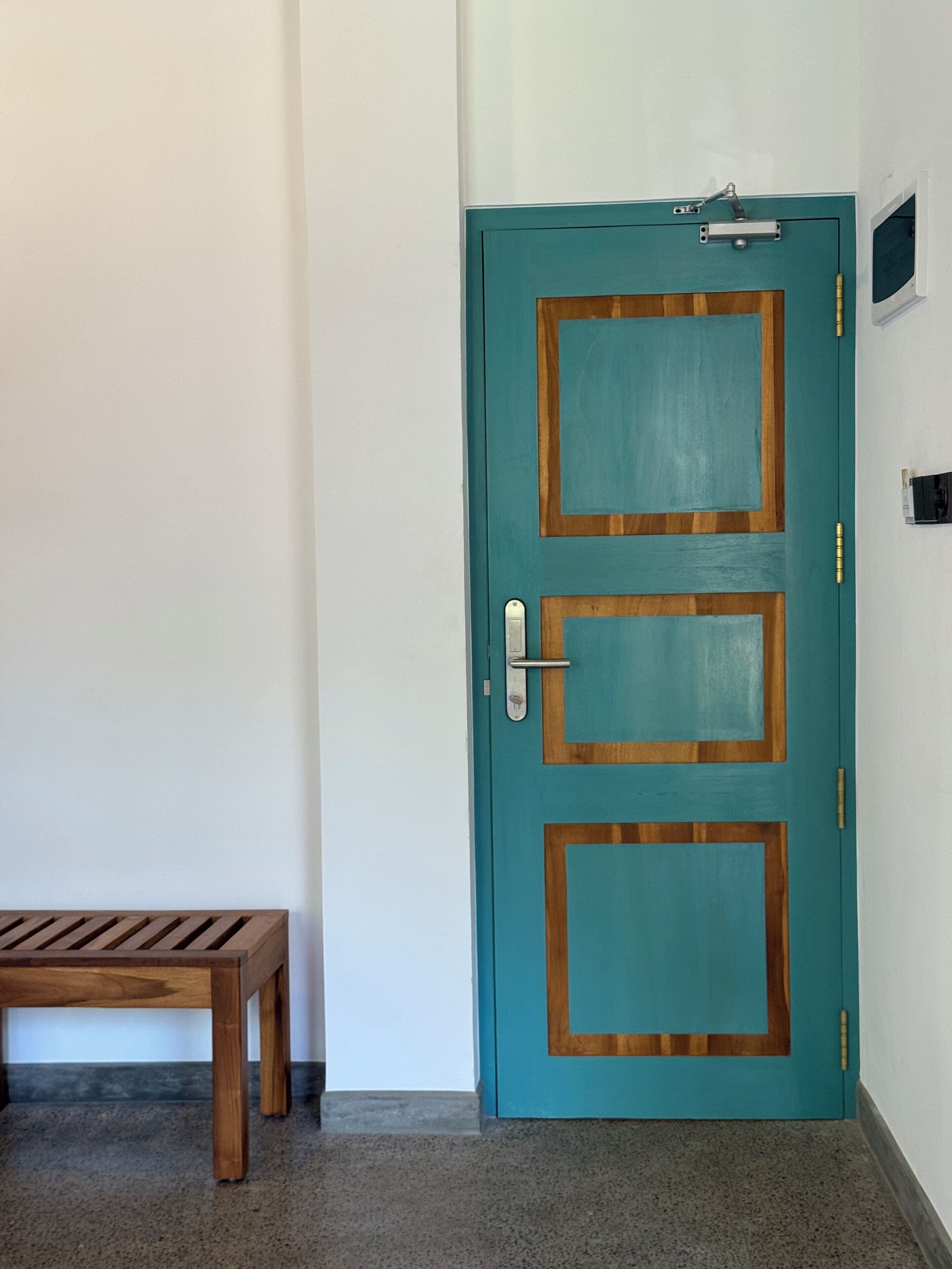 A teal door with a wooden frame and a metal handle, a bench, and a small window above the door.