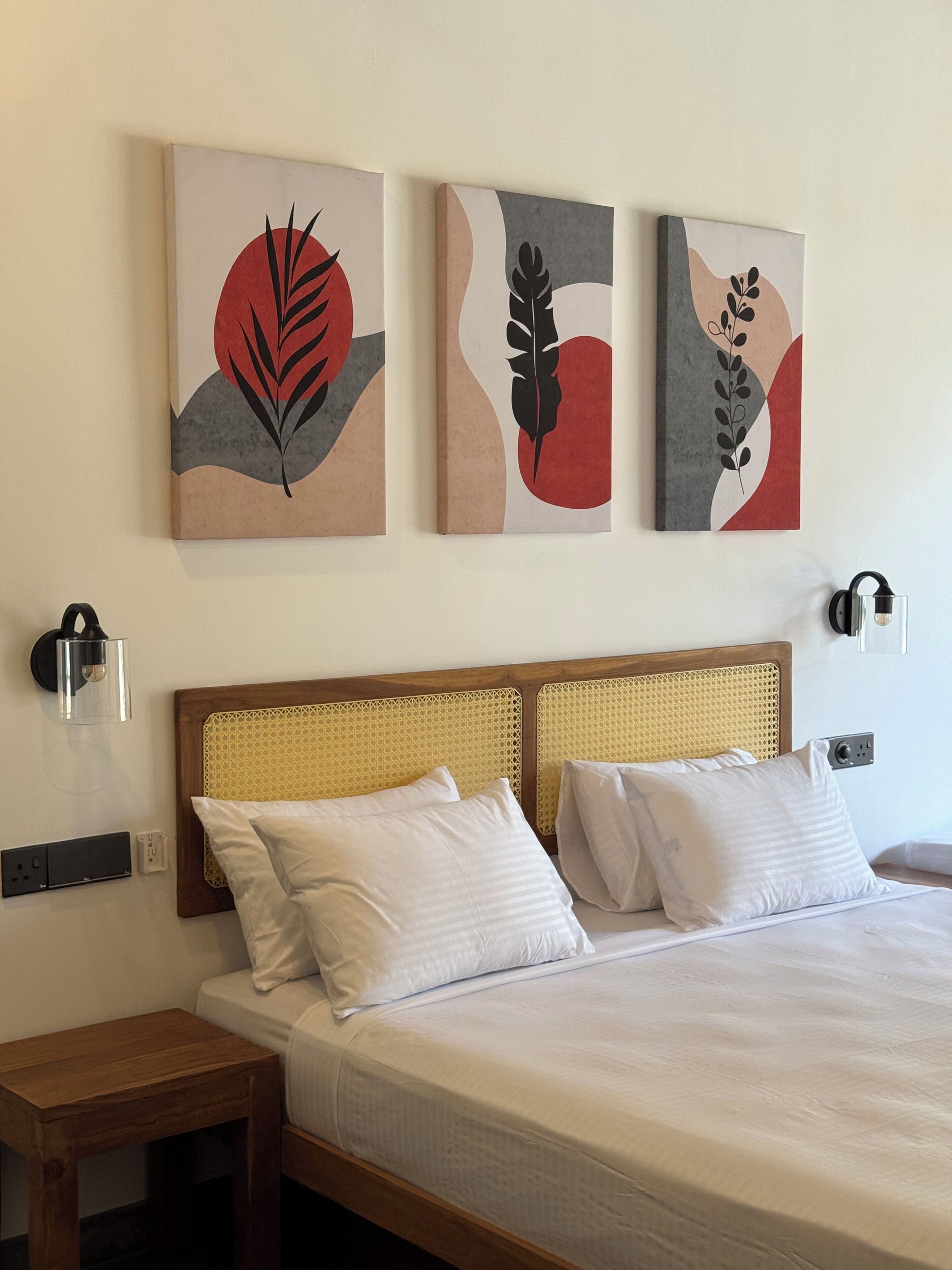 A hotel room with a wooden headboard, two white pillows, a bed with a white bedspread, and three abstract paintings on the wall.