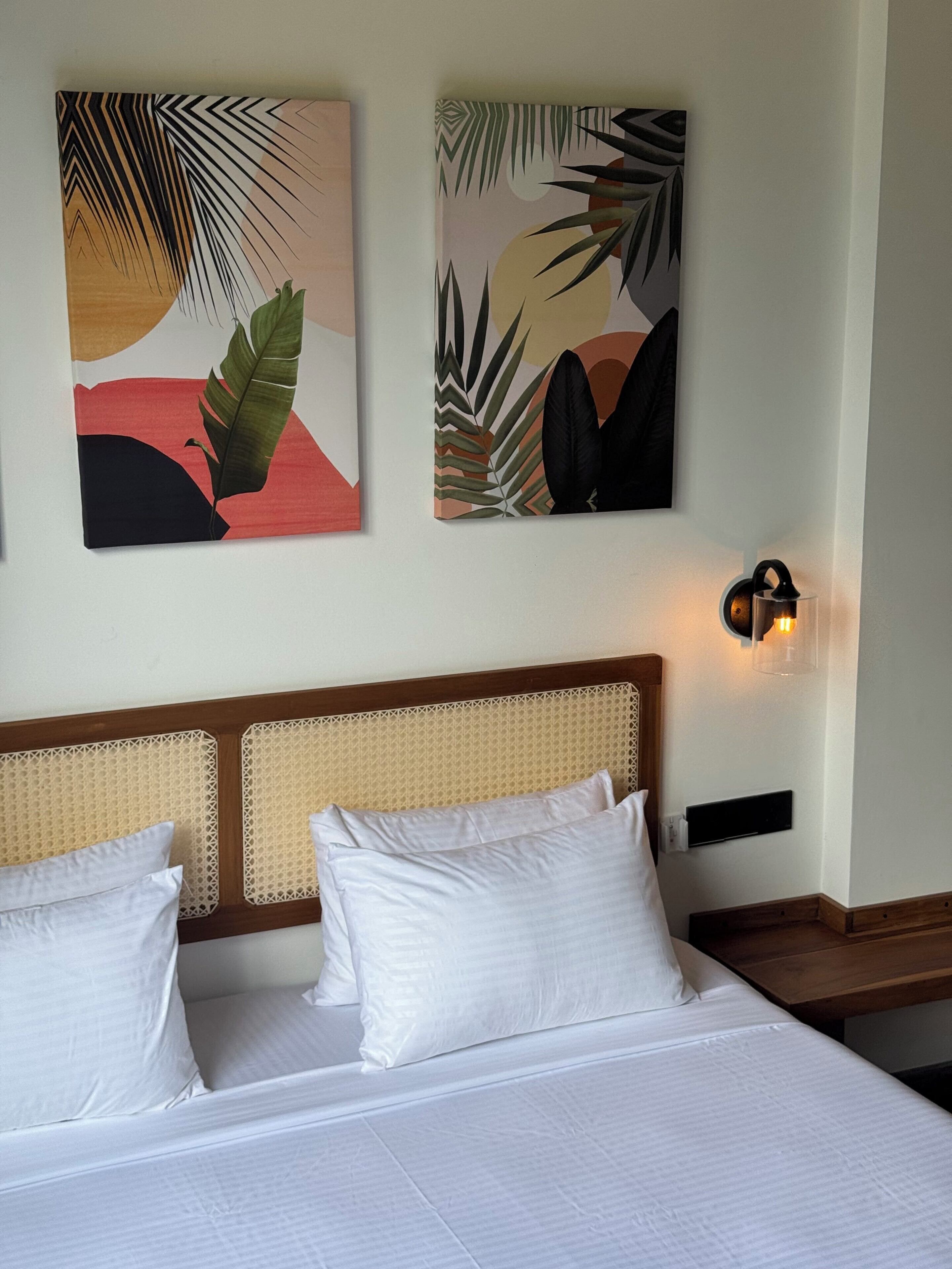 Ein Hotelzimmer mit einem Bett, weißen Kissen, einem gewobenen Kopfteil und zwei abstrakten Bildern an der Wand.