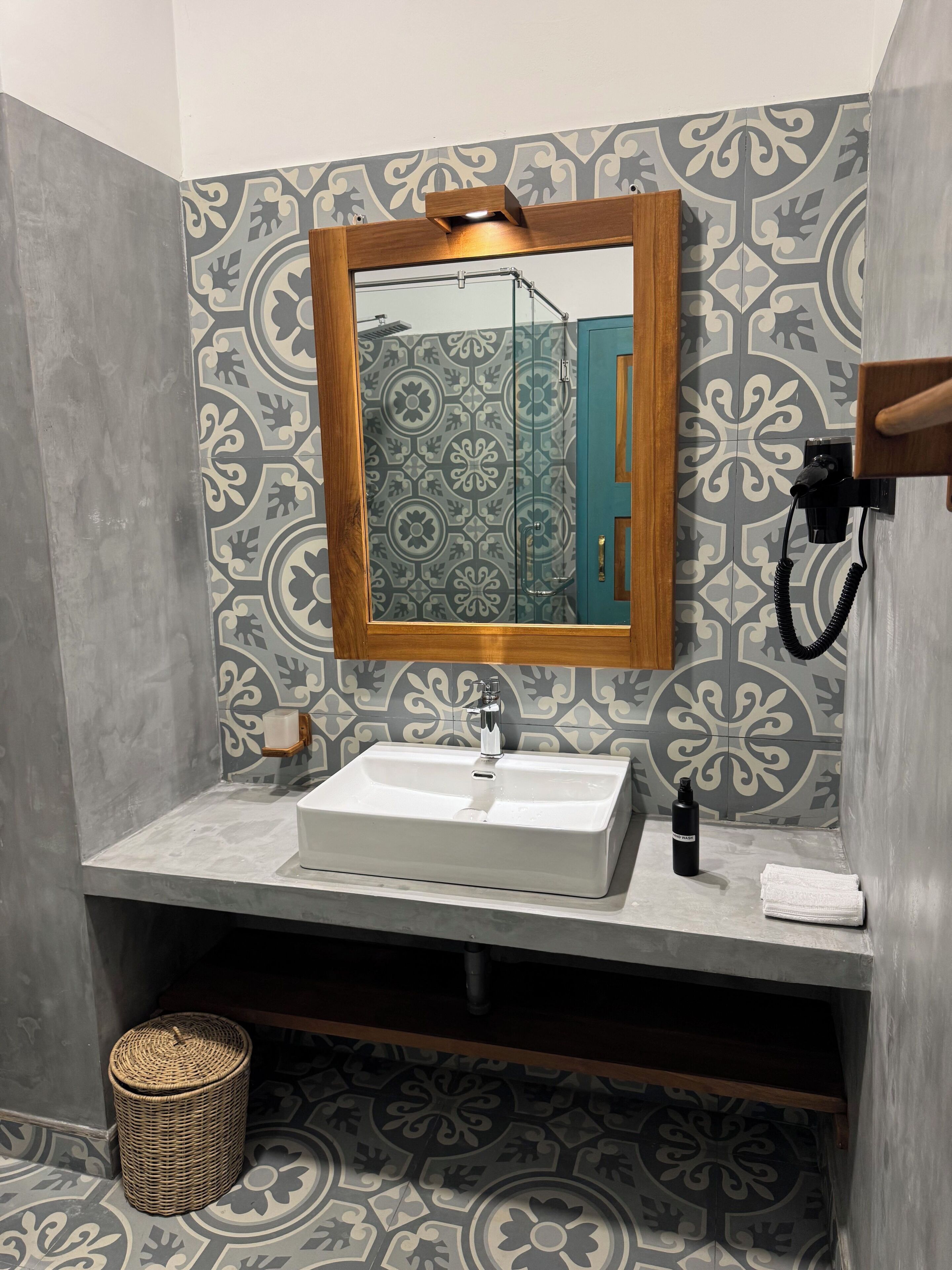 A bathroom with a white sink, a wooden-framed mirror, patterned wall tiles, a towel, and a small wicker basket.