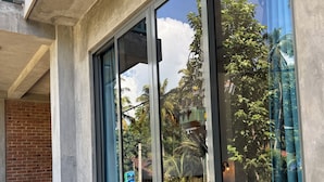Un edificio con grandes ventanales que reflejan palmeras y un cielo despejado. Hay macetas de concreto con plantas verdes a lo largo de la pared exterior.