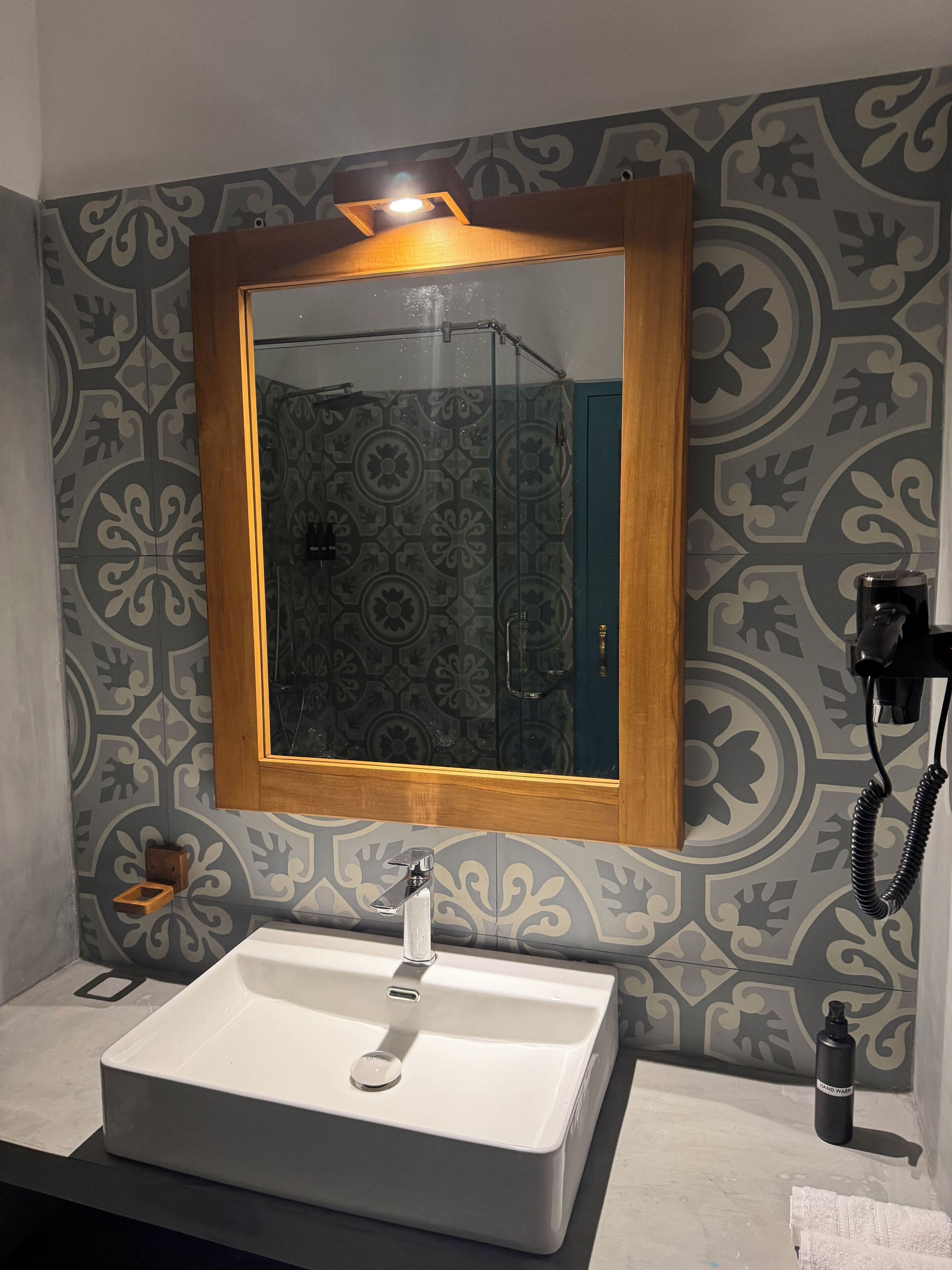 A bathroom with a white sink, a mirror with a wooden frame, patterned wall tiles, and a hand sanitizer bottle.