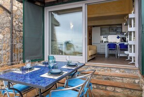 An outdoor dining area with a blue mosaic table, chairs, and a glass door leading to a kitchen with a sofa and blue chairs. - Stella Marina Apartment (Cala Piccola)