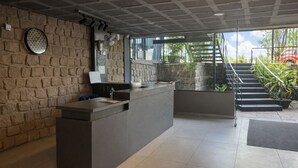 A reception area with a stone wall, a reception desk, a clock, and a staircase leading to an outdoor area with plants. - Rooms By DeVerveil (Mysore)