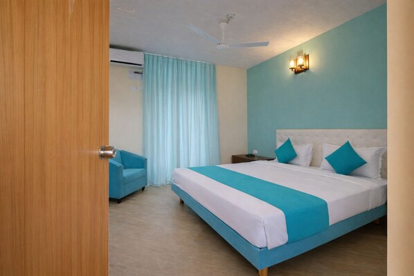 A hotel room with a bed, a chair, a nightstand, a ceiling fan, and an air conditioning unit. - Rooms By DeVerveil (Mysore)