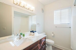 Baño con lavamanos blanco, espejo e inodoro. Hay toallas y artículos de aseo en la encimera.