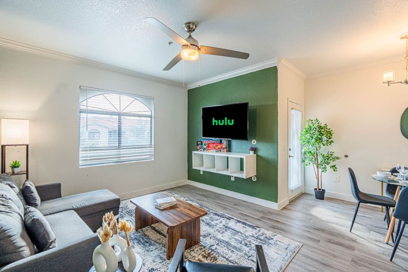 A living room with a ceiling fan, a television mounted on a green wall, a sofa, a coffee table, and a dining area with chairs.