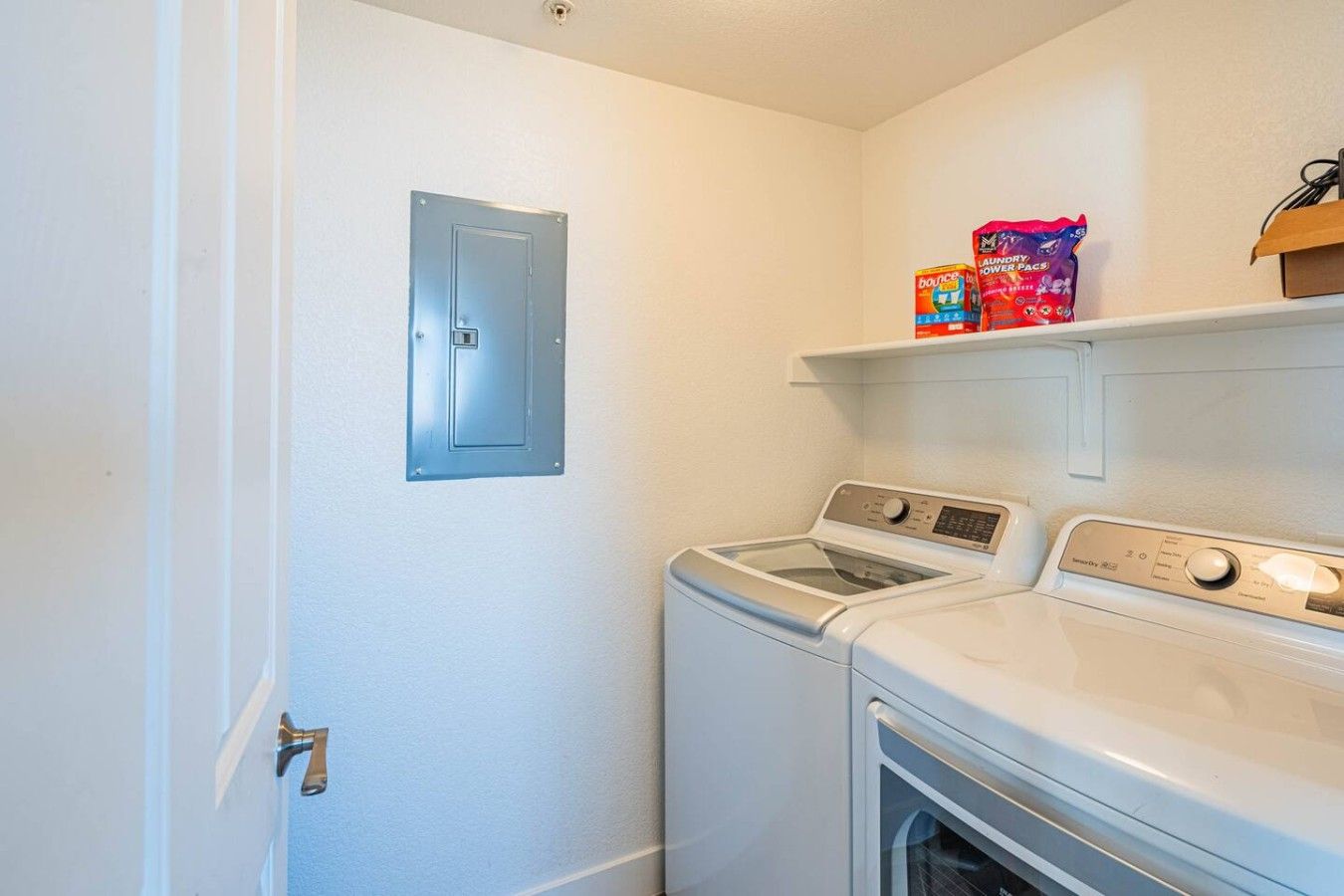 A laundry room with two front-loading washing machines, a dryer, and a shelf with laundry detergent.