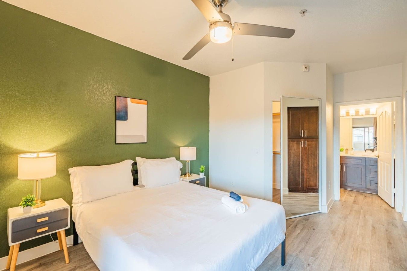 A bedroom with a bed, nightstands, a ceiling fan, and a view into a kitchen area.