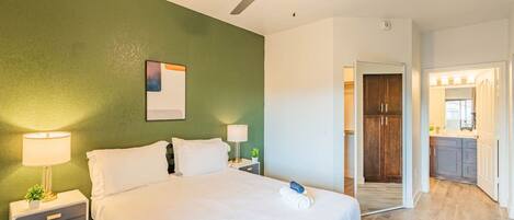 A bedroom with a bed, nightstands, a ceiling fan, and a view into a kitchen area.