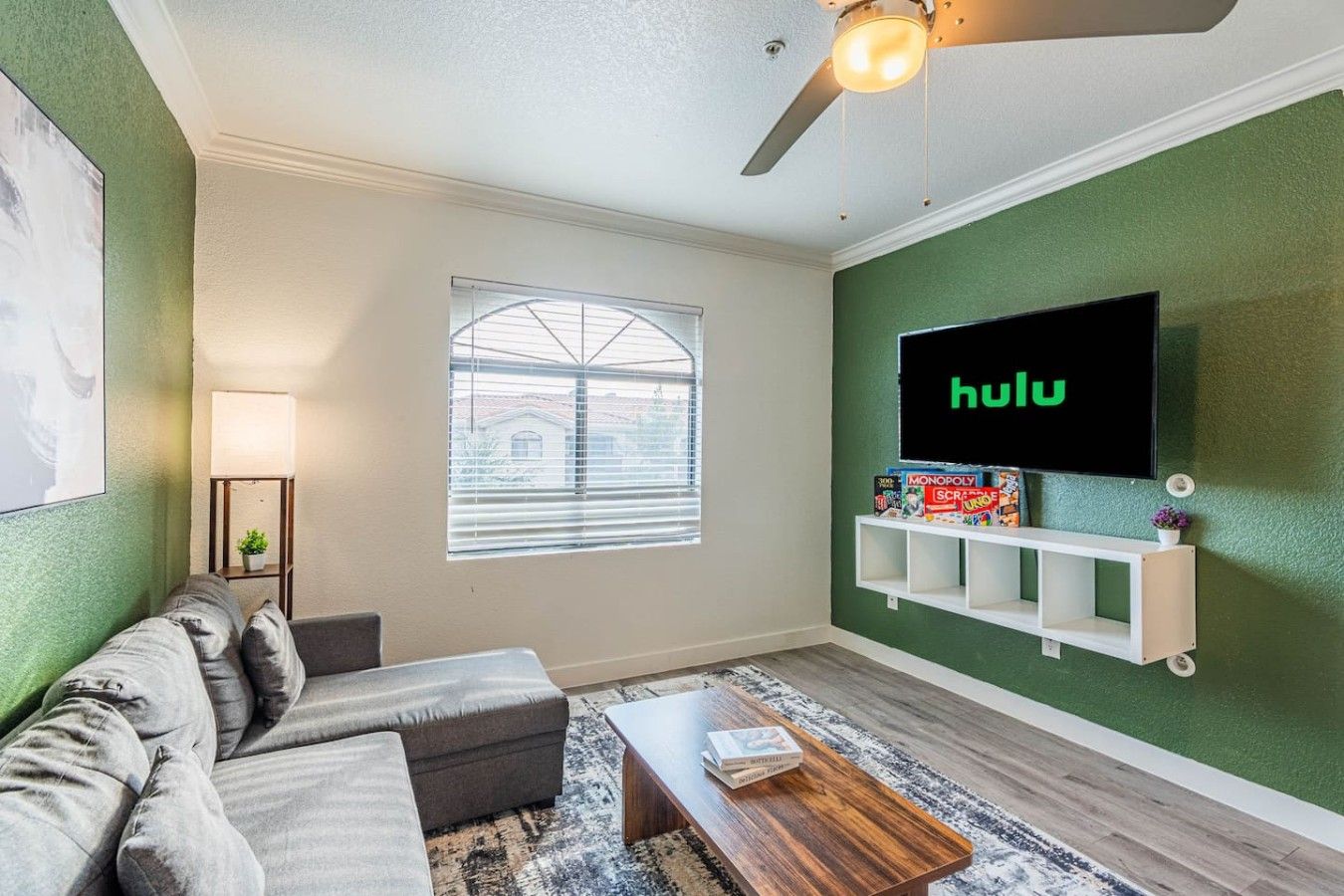 A living room with a sofa, a coffee table, a TV mounted on the wall, a window with blinds, and a ceiling fan.