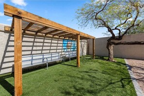 A wooden pergola structure with metal benches underneath, set against a wall with a mural, in a landscaped garden area with a tree and a clear sky. - 2BR-2BA Near ASU & Arizona Mills | Pool & W/D (Tempe)