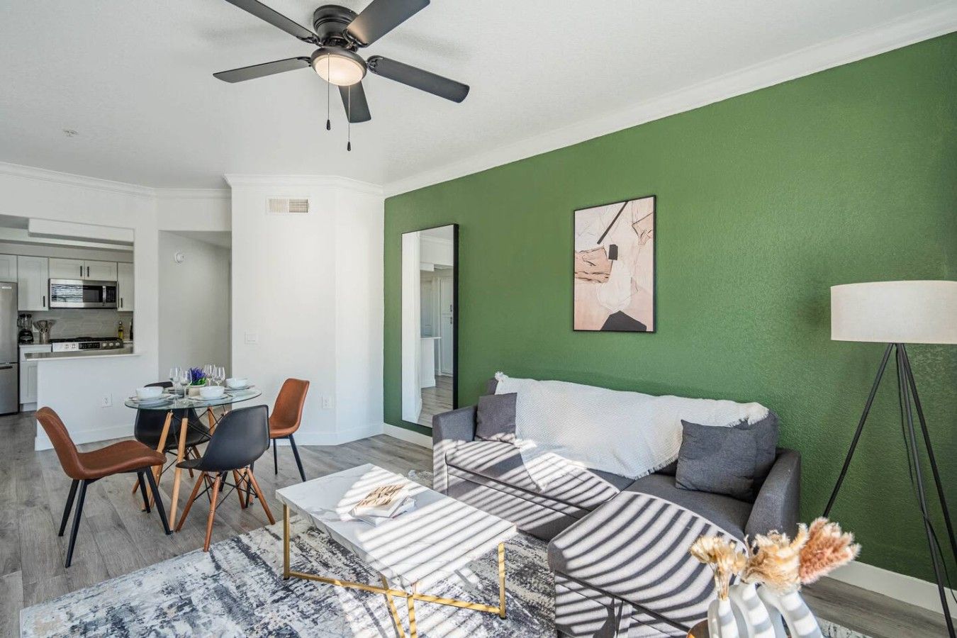 A modern living room with a green accent wall, a ceiling fan, a dining area with a glass table, and a kitchen in the background.