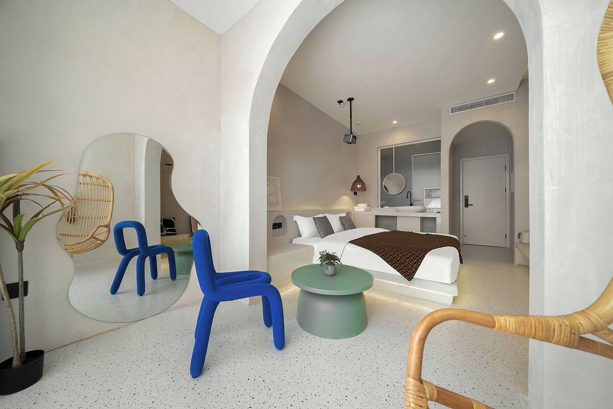 Une chambre d’hôtel moderne avec un grand lit, un coin salon comprenant un fauteuil et une petite table ronde, un grand miroir et une petite plante.