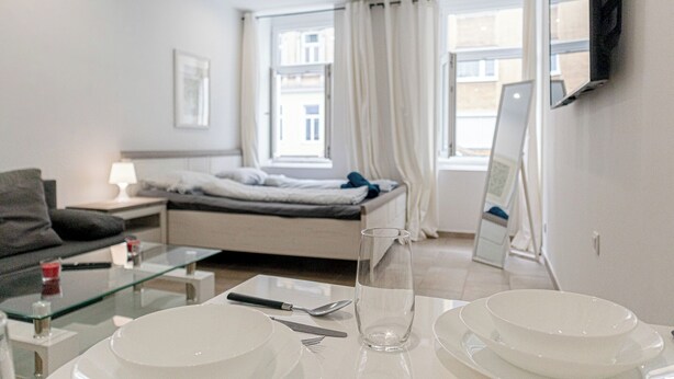 A modern hotel room with a bed, a glass coffee table, a dining table set with plates, a glass, and cutlery. - Central Vienna Elegant 1BR Apartment (Vienna)