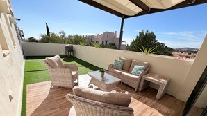 En udendørs terrasse med fletningsmøbler, et glastop bord og udsigt over det omkringliggende landskab.