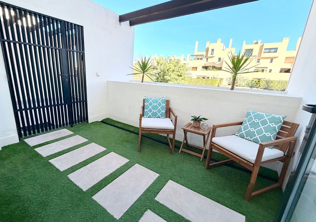 A balcony with artificial grass, two wooden chairs with cushions, a small table with a plant, and a black metal gate.