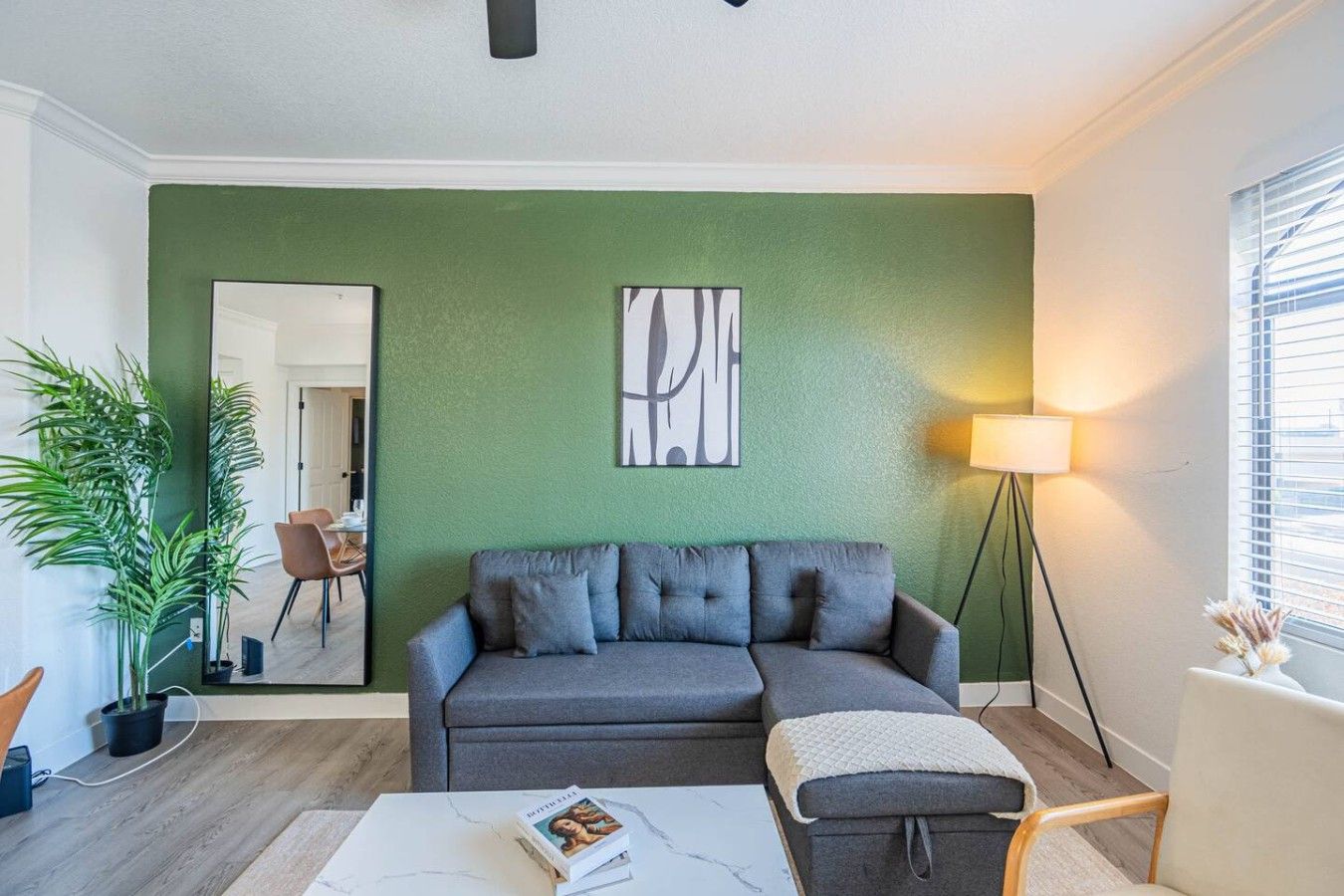 Una sala de estar con una pared verde, un sofá gris, una mesa de centro, una lámpara de pie y una planta en maceta.