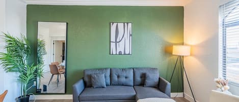 Una sala de estar con una pared verde, un sofá gris, una mesa de centro, una lámpara de pie y una planta en maceta.