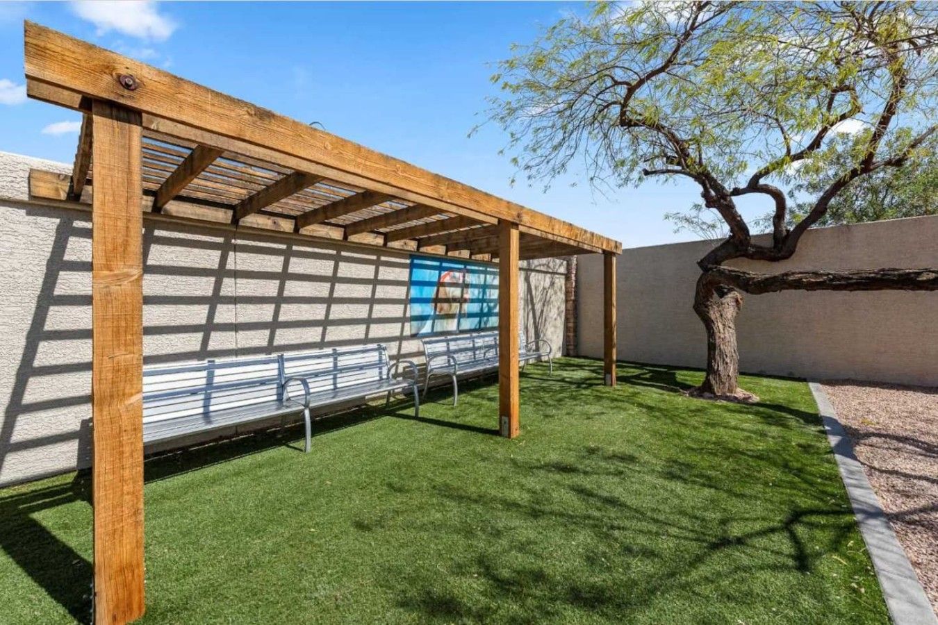 Une structure de pergola en bois, agrémentée de bancs en métal, une fresque murale et un arbre en arrière-plan.