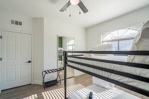 Une pièce avec des lits superposés, une porte, une fenêtre avec des stores et un ventilateur de plafond.