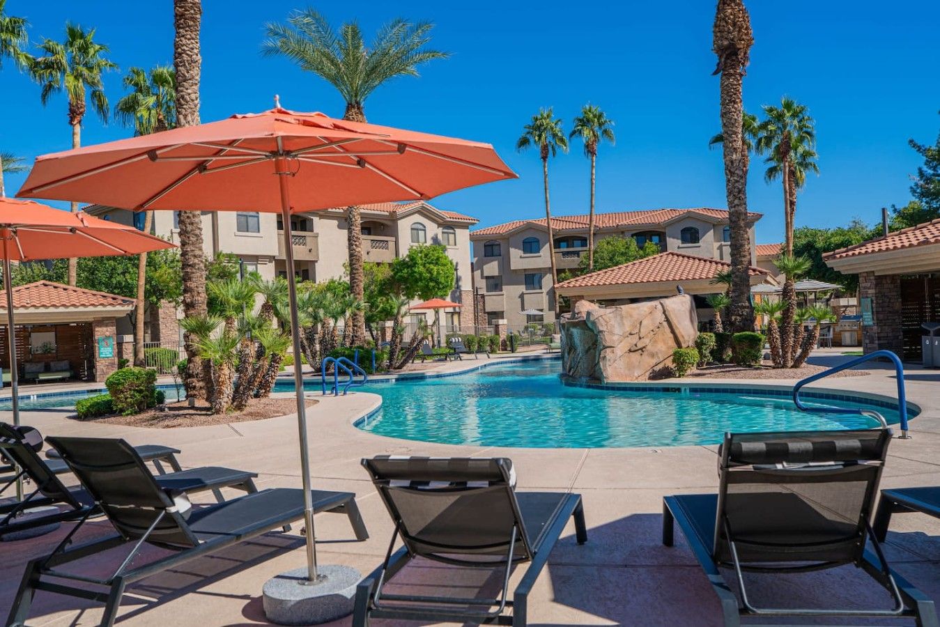 A pool area with lounge chairs and umbrellas, surrounded by palm trees and a building with a tiled roof.