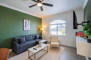 Un salon avec un mur vert foncé, un canapé gris, une table basse en bois, un lampadaire et une fenêtre avec des stores.