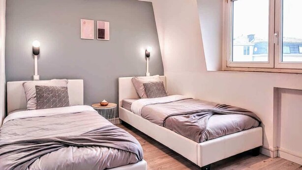 Un dormitorio con dos camas, una mesita de noche, una lámpara de pared y una ventana con vistas a edificios.