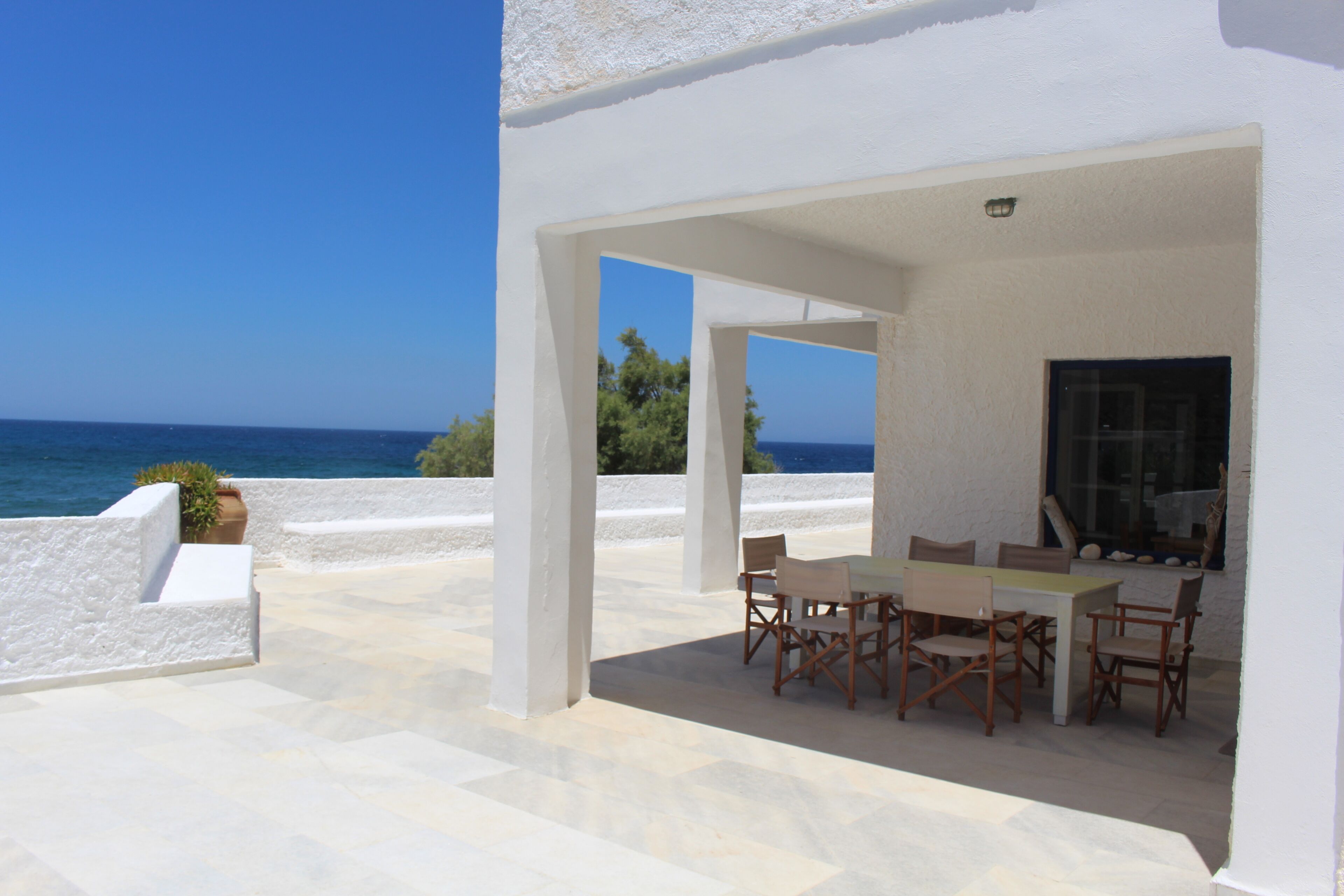 Um pátio externo com mesa e cadeiras, uma parede branca e vista para o mar.
