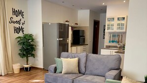En moderne stue med en grå sofa, en grøn sofa og et sofabord. I baggrunden er der et køkkenområde med et køleskab og skabe. En plante står på gulvet ved siden af sofaen.