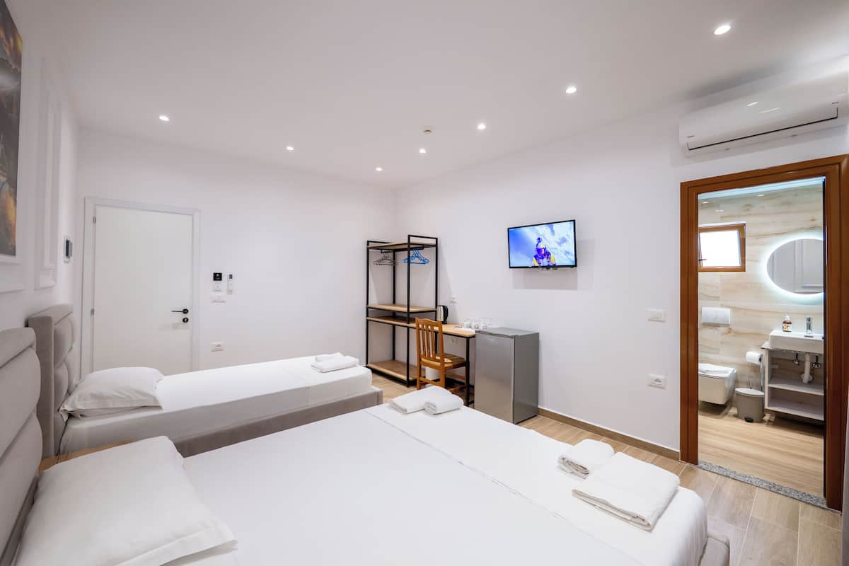 Camera d'albergo con due letti, una scrivania con sedia, una televisione, uno specchio e bagno parzialmente visibile da una porta aperta.