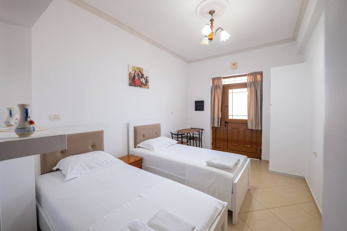 Una camera d'albergo con due letti singoli, un tavolino, una sedia, un lampadario a soffitto e una porta con tende.