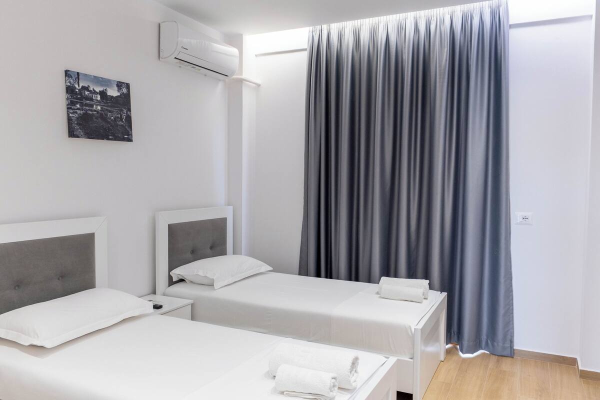 Una camera d'albergo con due letti, un condizionatore fissato al muro, una tenda grigia e un quadro incorniciato appeso al muro.