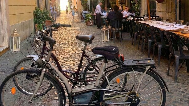 A cobblestone street with a bicycle parked in the foreground. There are outdoor tables and chairs, and buildings with balconies and plants. - Navona Apartment (Rome)
