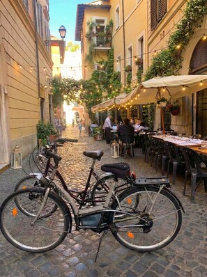 En brostensbelagt gade med en cykel parkeret i forgrunden. Udendørs borde og stole, samt bygninger med altaner og beplantning.