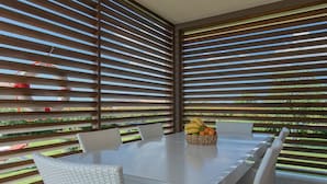 A dining area with a white table and wicker chairs, a fruit basket, and a view through horizontal blinds. - Modern villa with pool, garden and sea view for 8 people in Istria (Galižana)