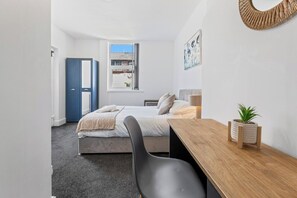 Ein Schlafzimmer mit Bett, Schreibtisch, Stuhl, Zimmerpflanze, Tür und Fenster.