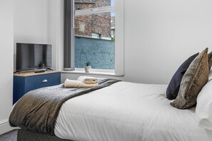 Ett sovrum med en säng, en tv på ett stativ, ett fönster med utsikt över en tegelvägg och en liten växt på fönsterbrädan.