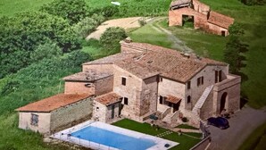 Une vue aérienne d’un grand bâtiment rustique en pierre, doté d’une piscine et entouré d’une végétation luxuriante.