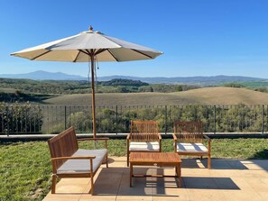A patio with wooden chairs and a table, an umbrella, and a scenic view of rolling hills and mountains.