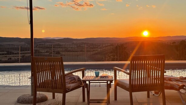 Une vue sur le coucher de soleil depuis une terrasse, avec deux chaises en bois, une table et un parasol.