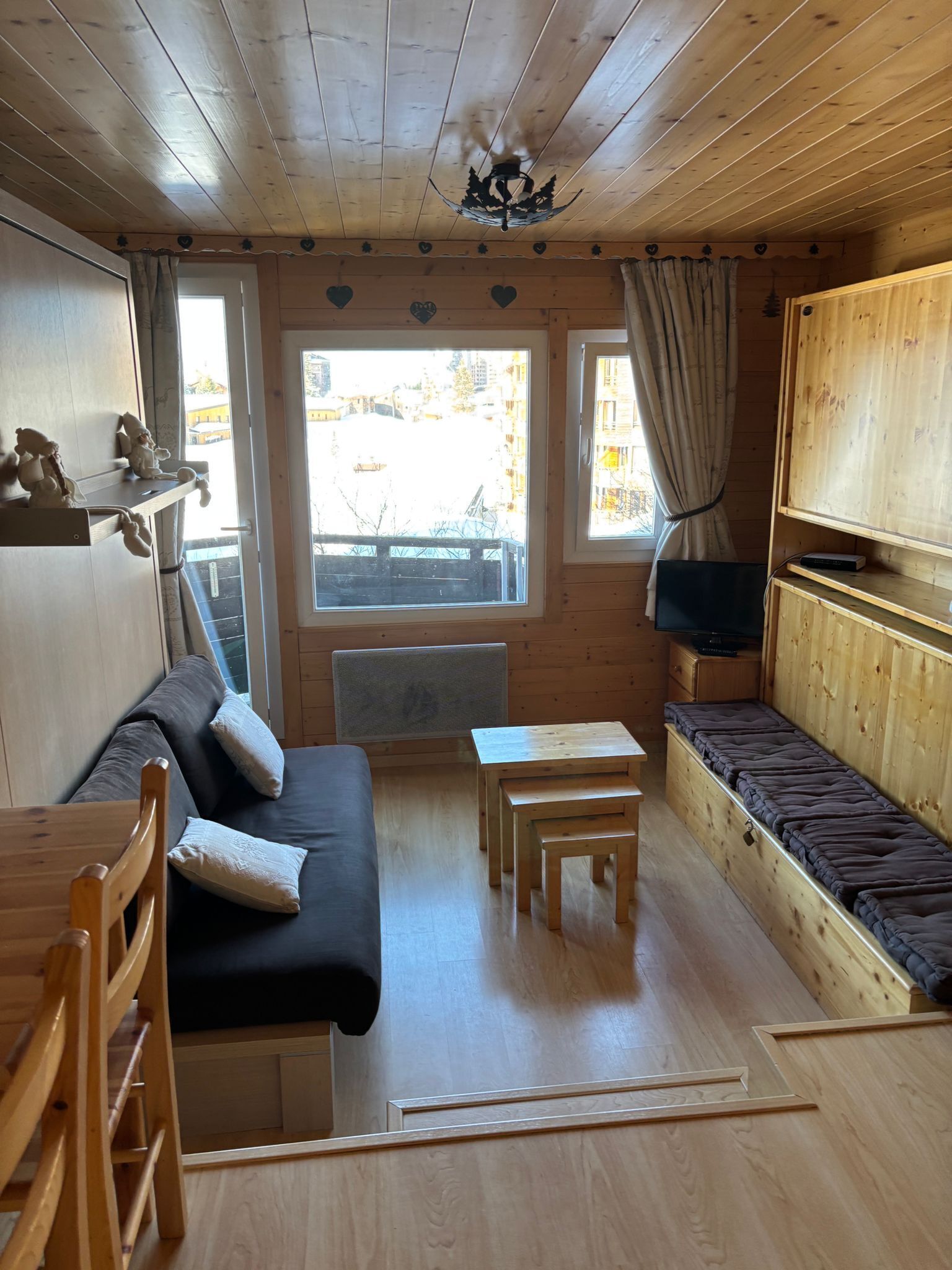 A cozy cabin interior with a wooden ceiling, a sofa, a small table, a TV, and a window with curtains.