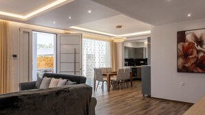 Ein modernes Wohnzimmer mit Sofa, Esstisch, Stühlen und einer Küche im Hintergrund. Ein großes Fenster mit Vorhängen.