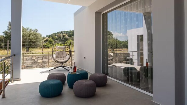 Ein moderner Balkon mit einer Hängematte, runden Hocker und einer Glastür, die zu einem Außenbereich mit Blick auf Bäume und einen Zaun führt.