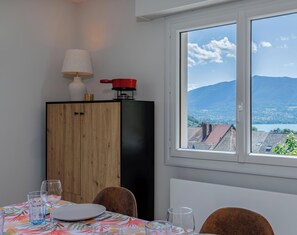 Ein Essbereich mit einem Tisch für zwei Personen, einem Schrank mit einem roten Topf darauf, einer Lampe und einem Fenster mit Blick auf Berge und einen See.