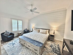 Ein Schlafzimmer mit einem großen Bett, zwei Nachttischen mit Lampen, einem Sessel, einem Deckenventilator und einem Fenster mit Jalousien.