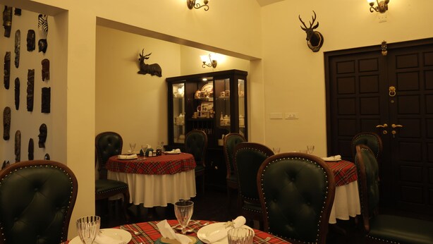 A dining room with four tables, each with a red and white checkered tablecloth, surrounded by green upholstered chairs. There are wall-mounted light fixtures and a door with a decorative antler head. - Lynton Resorts (Kotagiri)