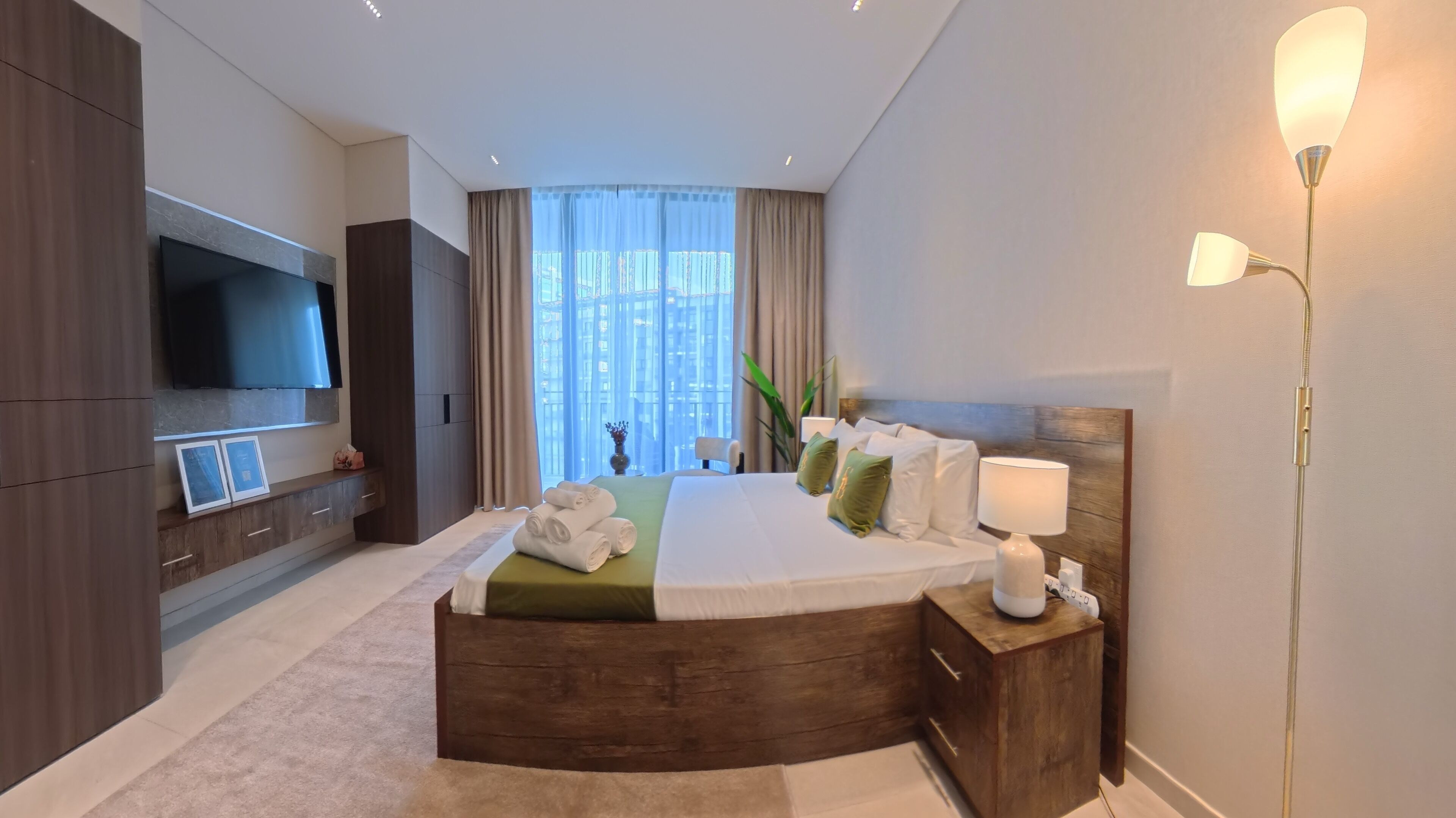 Une chambre d’hôtel moderne dotée d’un lit aux lignes courbes, d’un téléviseur à écran plat, d’une table de chevet avec une lampe et offrant une vue sur la ville grâce à de grandes fenêtres.
